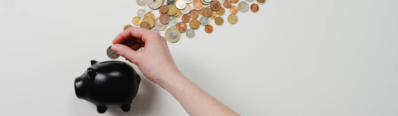 Foto de uma mão colocando moedas em um porquinho de porcelana preto, representando a economia de dinheiro.