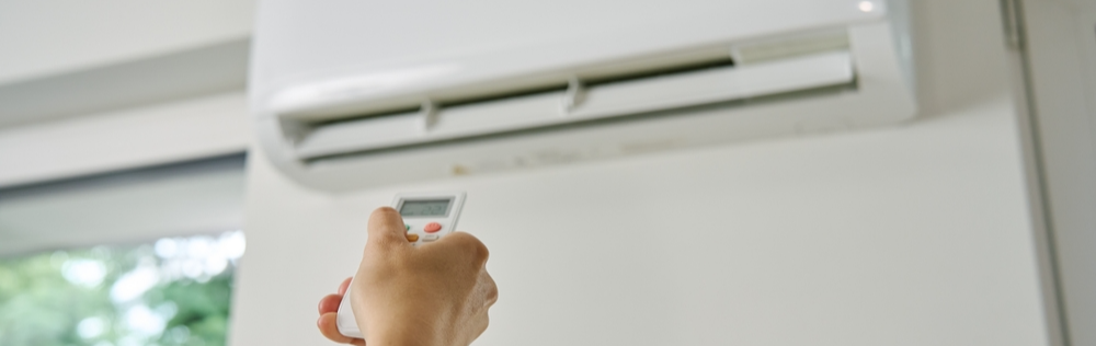 Uma mão segura o controle remoto de um ar-condicionado, ajustando a temperatura para 26°C. Ao fundo, o aparelho está preso em uma parede de cor branca.